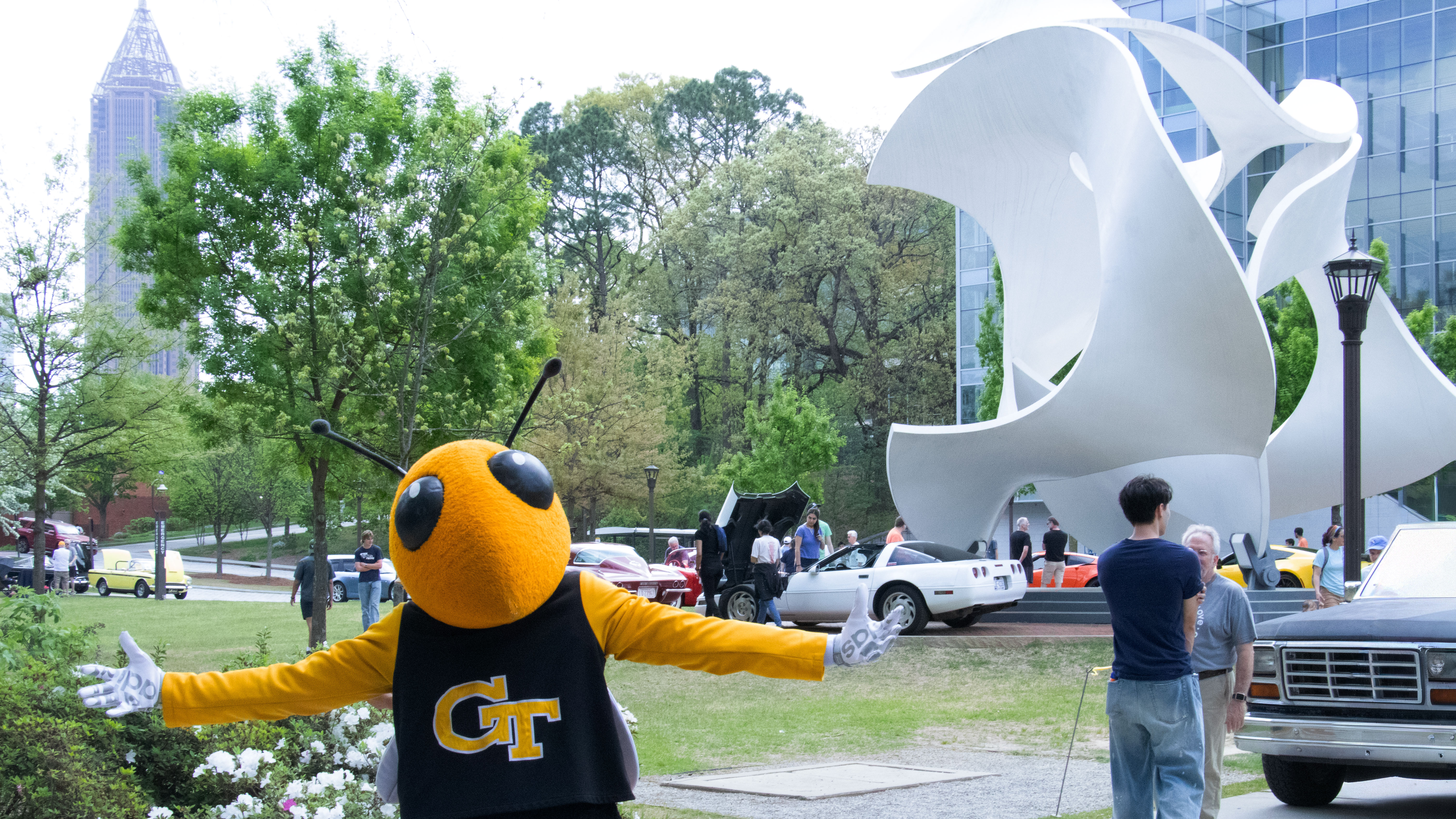 Georgia Tech's Buzz mascot stands with arms outstretched on Tech Green near the large white Kendeda Building sculpture, with classic cars and visitors in the background.