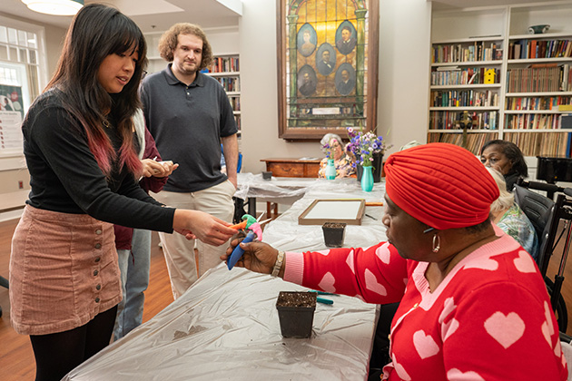 Two people exchange gardening tools across a table prepared for a planting activity in a room with bookshelves and potted plants.