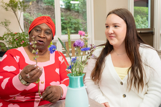 Two people sit beside a vase of flowers while one holds a flower stem during a floral arrangement activity indoors.