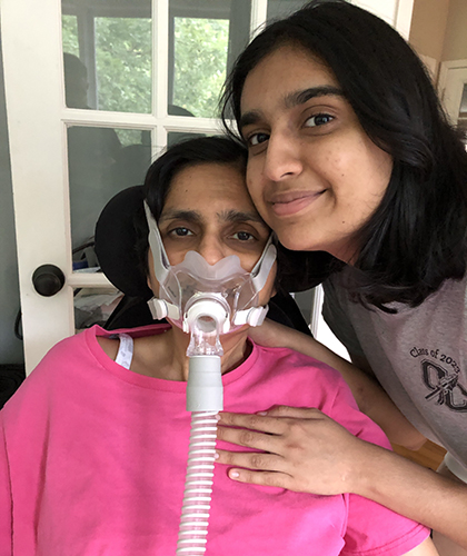Mother wearing a noninvasive ventilation mask sits with her daughter, who rests a hand on her shoulder, as they pose for a photo.