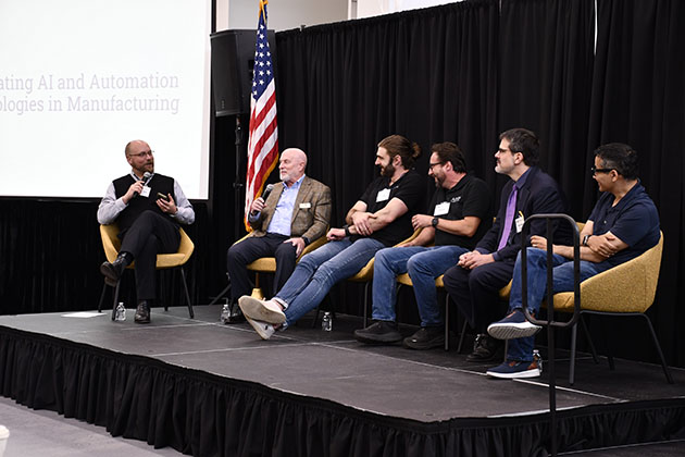 Panel discussion on stage with six speakers seated in chairs, holding microphones, with a U.S. flag and presentation screen behind them.