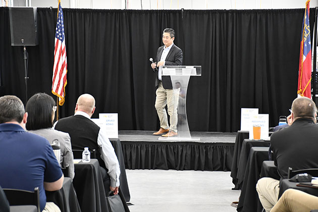 Speaker standing at a podium addressing an audience, with a U.S. flag and black curtain backdrop in a conference setting.