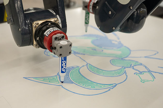 Robotic arms using marker tools to draw a detailed green and blue pattern on a flat surface.