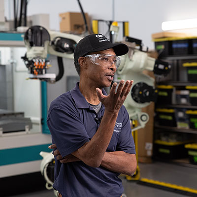 Individual wearing safety glasses and a cap speaking in a lab environment with robotic equipment in the background.

