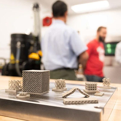 Close-up of metal 3D-printed lattice samples on a table, with people and manufacturing equipment blurred in the background.
