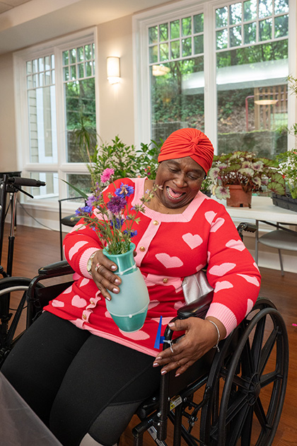 Person seated in a wheelchair arranges a bouquet of flowers in a vase inside a bright room with windows and potted plants.
