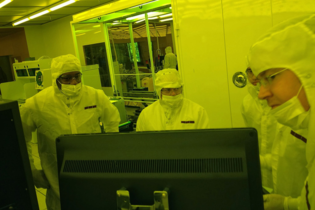 Three people in full cleanroom suits stand around a computer monitor in a yellow‑lit lab, with one person gesturing toward the screen.