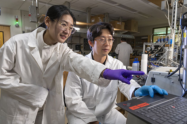 female researcher wearing a purple glove and q white lab coat pointing at a screen in a lab while a male researchers who is sitting looks on