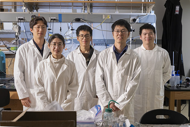 group of five researchers wearing white lab coats posing in front of standard lab equipment