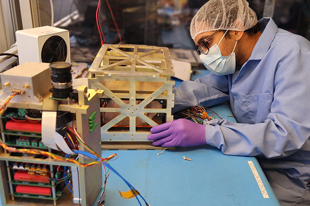 Technician wearing protective clothing and purple gloves assembling a metal cube-shaped satellite component on a workbench with electronic equipment and wiring nearby.