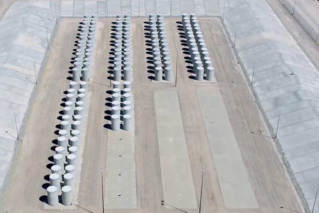 Aerial view of a nuclear waste storage facility with rows of large cylindrical concrete containers arranged in parallel lines on a flat, secured site.
