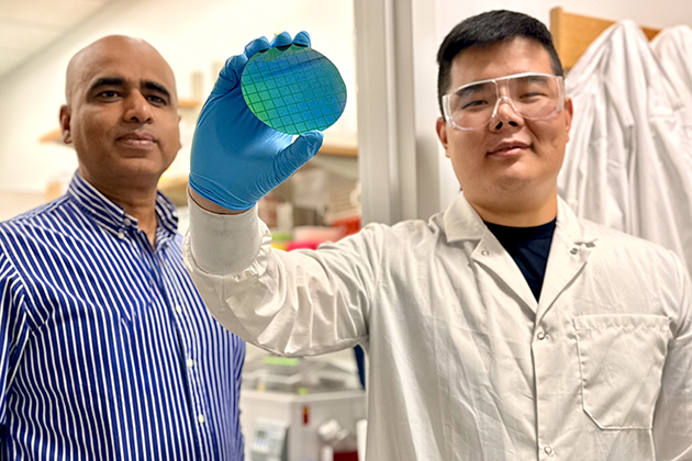 Two researchers in a laboratory examine a blue-green silicon wafer. One, wearing a lab coat, safety goggles, and a blue glove, holds the wafer up for inspection while the other stands beside them. Laboratory equipment is visible in the background.