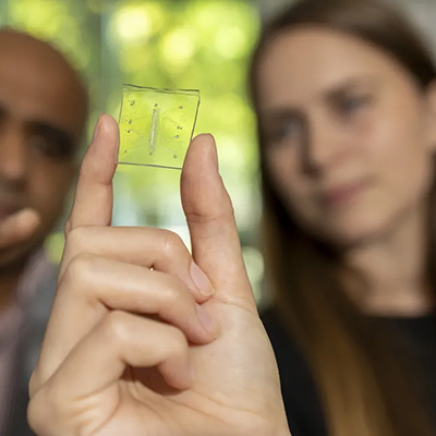 Researcher holds a small transparent microdevice between two fingers while collaborators observe in the background.