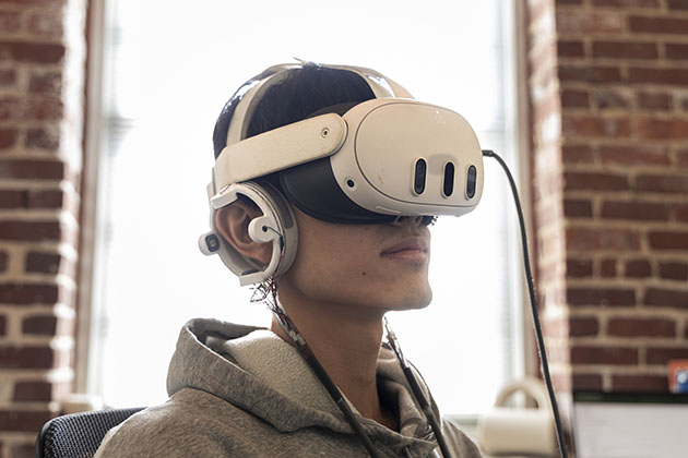 Person wearing a virtual reality headset and over-ear headphones, seated indoors near a window with exposed brick walls in the background.