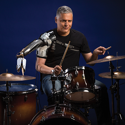 Person playing a drum set while using a robotic prosthetic arm, demonstrating advanced assistive technology and brain-controlled prosthetics.