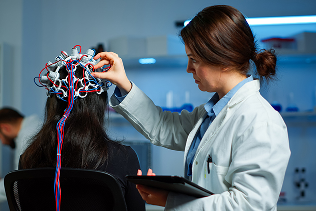 Researcher fits and adjusts an EEG brain-monitoring cap with electrode leads on a seated participant in a neuroscience laboratory while holding a tablet.