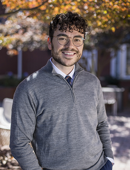 Person dressed in a gray quarter-zip sweater over a collared shirt and tie, standing outdoors with autumn foliage and brick buildings in the background