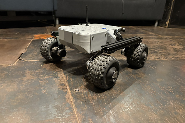 A small four-wheeled robotic rover with rugged tires sits on a metal floor indoors. The rover has a gray rectangular body, antennas, and mounted components designed for mobility and testing.