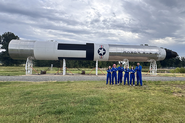 A large black-and-white rocket displayed horizontally on support stands in an outdoor grassy field. Six people wearing matching blue flight suits stand in front of the rocket under a cloudy sky.