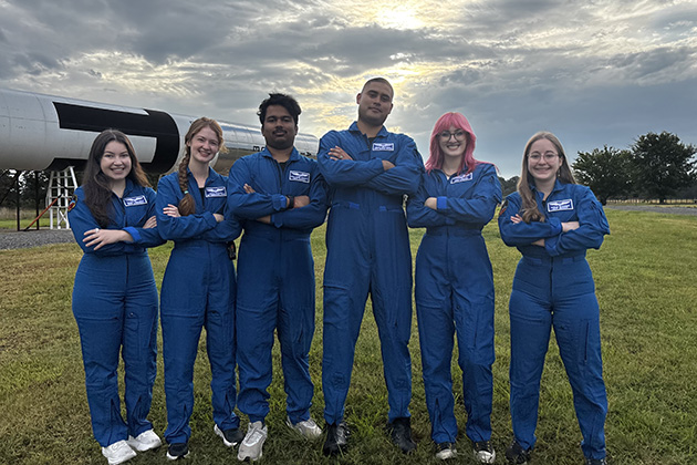 Six people wearing matching blue flight suits stand with arms crossed in a grassy field beside a large rocket display. The group poses in front of the rocket under a cloudy sky with sunlight visible through the clouds.