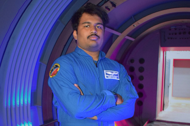 A person wearing a blue flight suit stands with arms crossed inside a curved, tunnel-like structure illuminated with blue and purple lighting. The corridor extends into the background, creating the appearance of a spacecraft or space simulation environment.