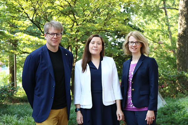 Three individuals standing outdoors in a landscaped area with green trees and shrubs in the background.