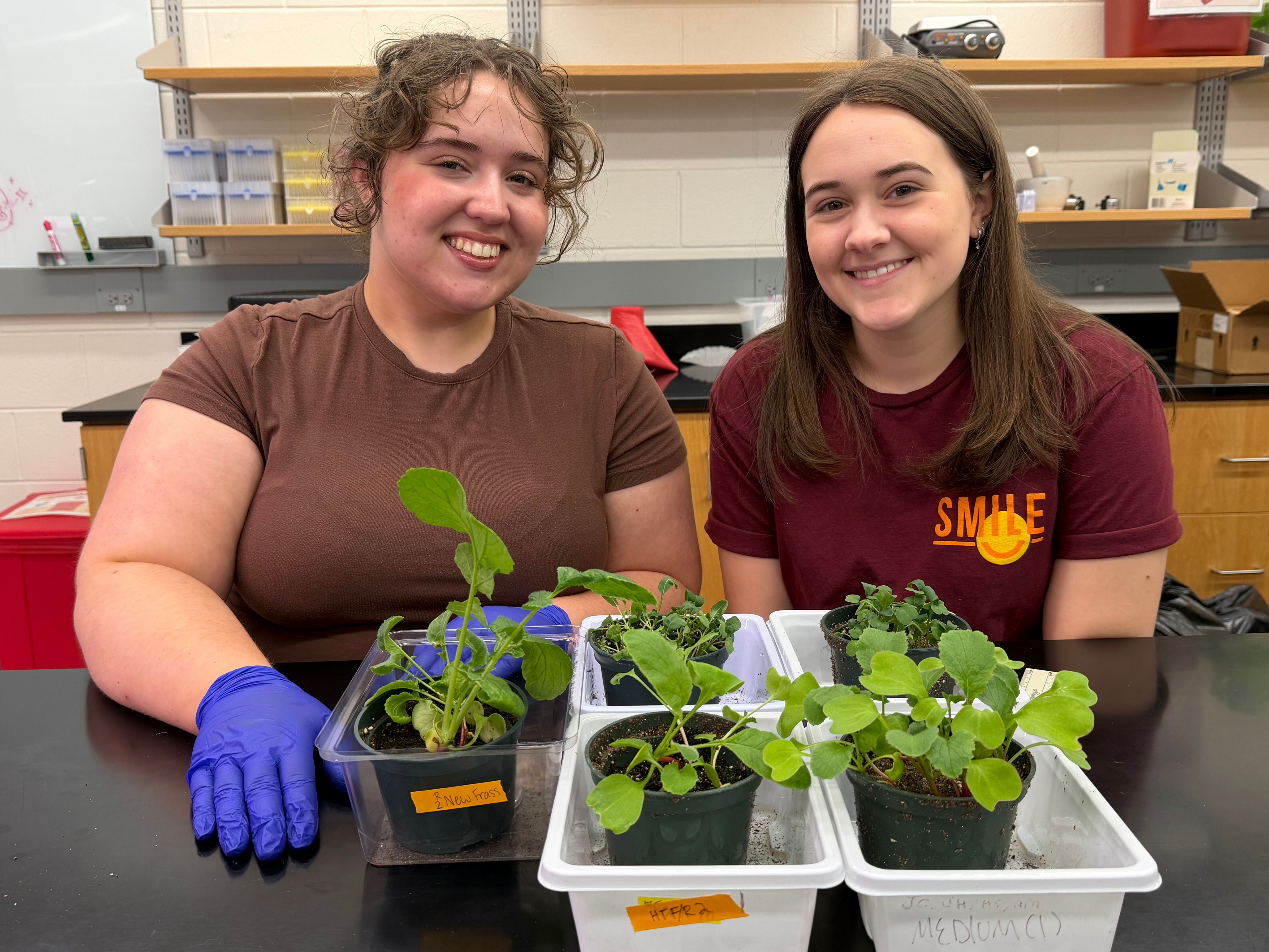 Two female biology students smile behind several small plants in plastic containers.