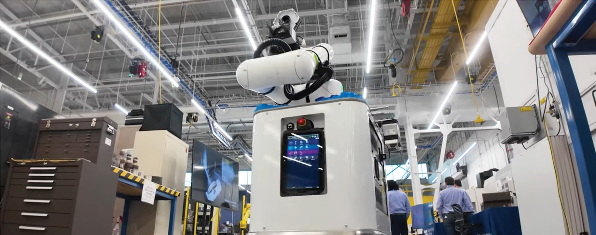 Mobile robotic system with a mounted robotic arm and onboard screen operating inside a bright manufacturing lab with equipment, workstations, and overhead lighting visible.