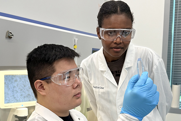 Two researchers wearing safety goggles and white lab coats examine a small rectangular semiconductor device in a laboratory. One researcher holds the device with a blue nitrile glove while the other closely observes it. Laboratory equipment and a microscope monitor are visible in the background.