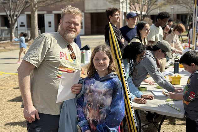 A little girl with her father standing outside with other people sitting down at a table in the background.