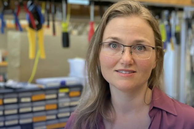 Researcher wearing glasses and a purple blouse stands in a laboratory with tools, equipment, and storage drawers visible in the background.