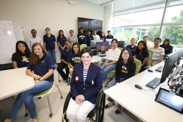 Group of Georgia Tech students and faculty gathered in a bright computer lab with workstations, including a wheelchair user seated in the front row.