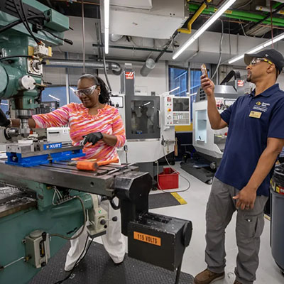Two people work beside machining equipment in a laboratory, with one operating the machine while the other holds up a device.