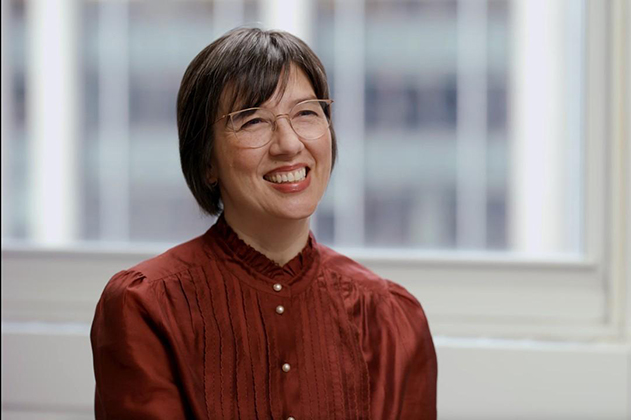 Portrait of a researcher seated indoors in front of large windows, wearing glasses and a burgundy blouse, photographed in an office or academic setting.Provide your feedback on BizChat
