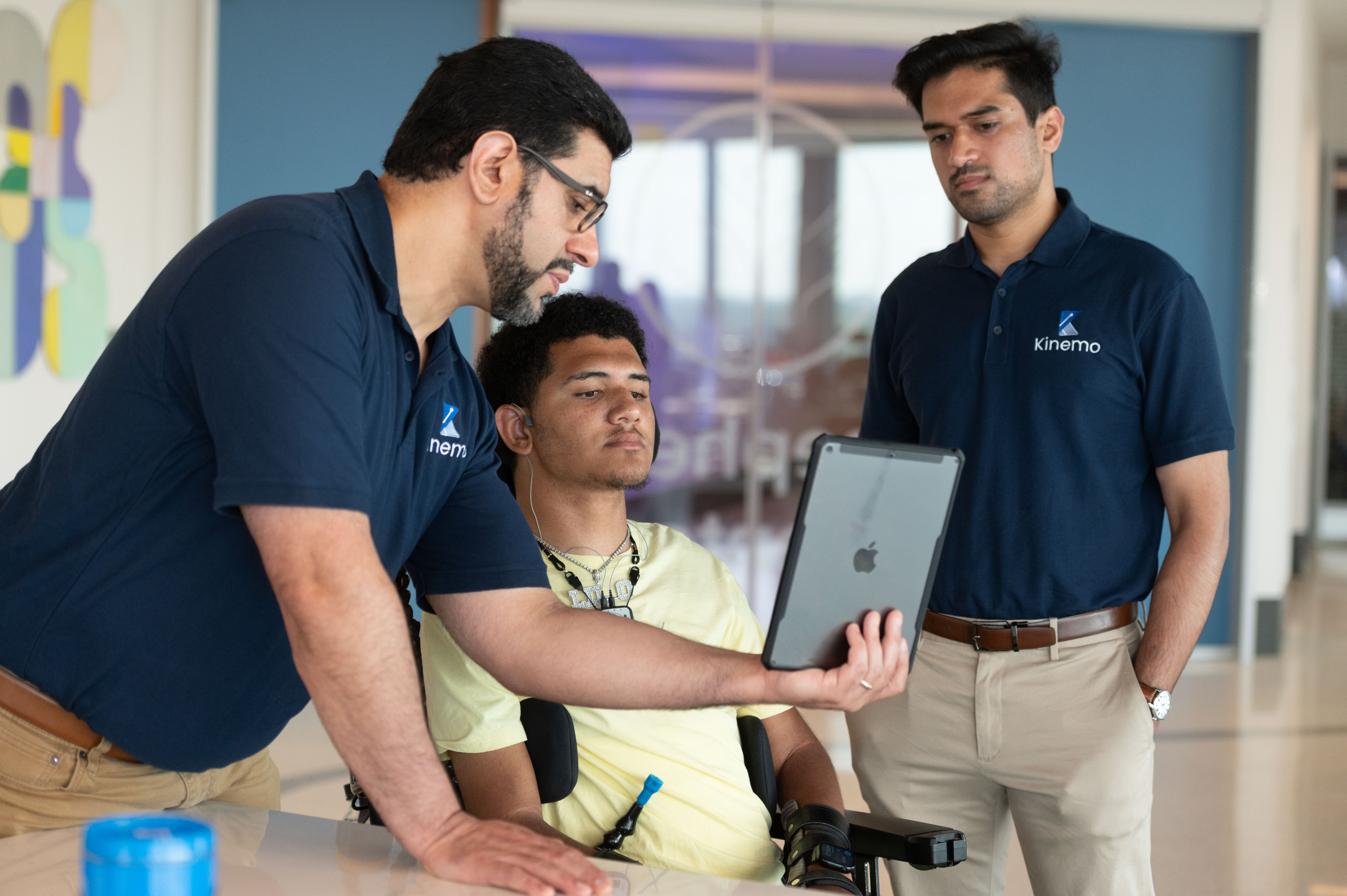 Three people review a tablet together in a lab setting, with one seated individual using a powered wheelchair and assistive device.