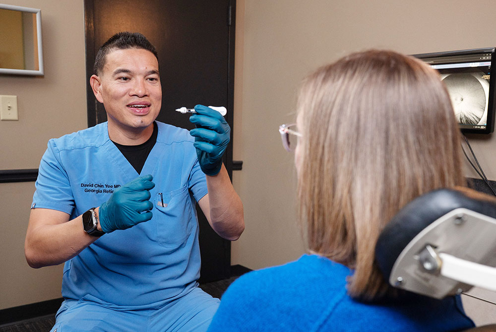 Person in blue medical scrubs demonstrates a small device to a seated patient in a clinical exam room with medical equipment visible in the background.