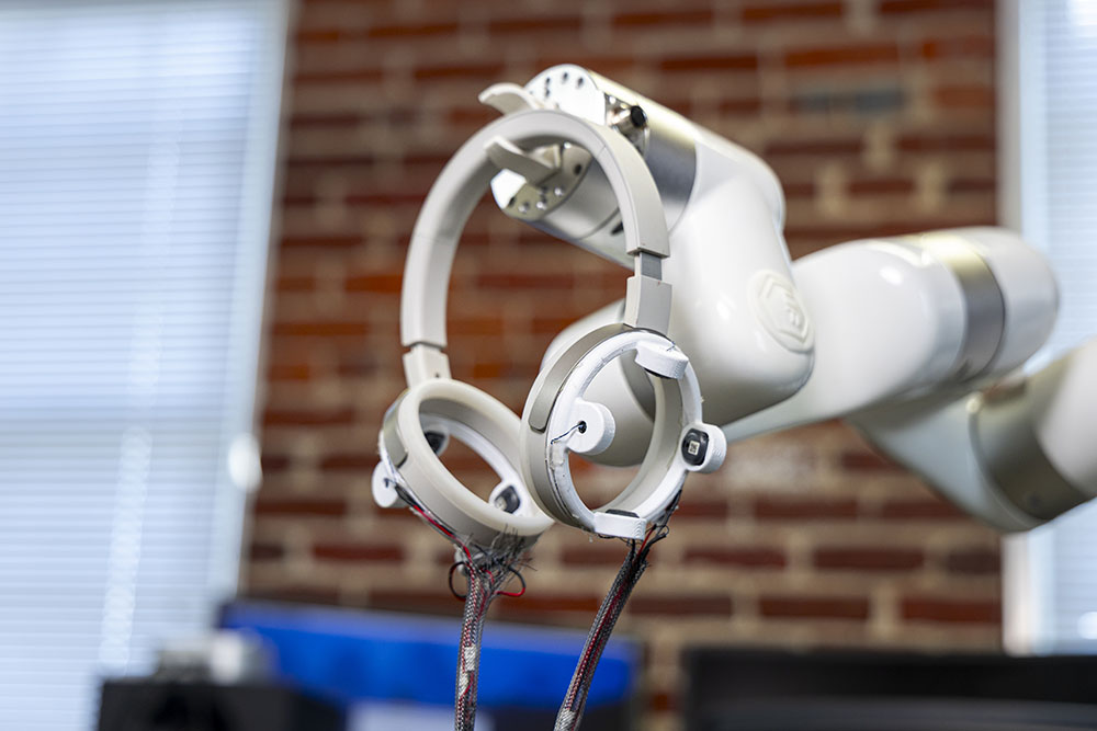 Robotic arm holding circular sensor devices with exposed wiring in a lab setting with a blurred brick wall background.