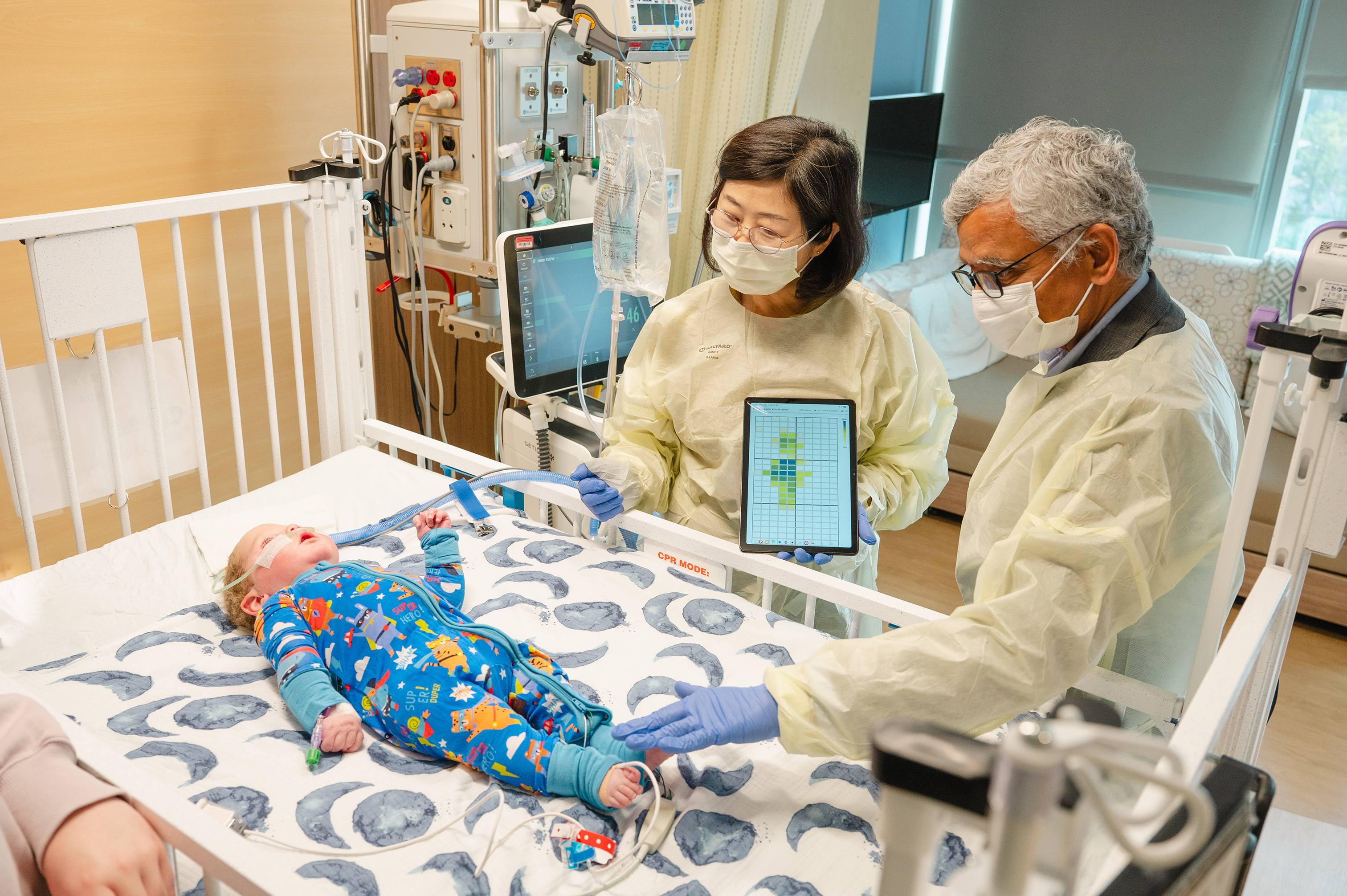 Two adults wearing protective gowns and gloves stand beside a hospital crib, using a tablet device while examining an infant lying on the mattress as medical equipment and monitors surround the crib.