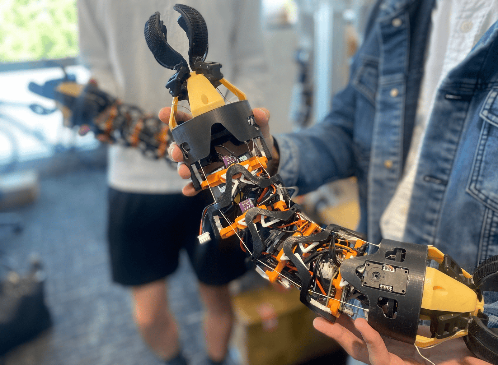 Close-up of a modular, snake-like robotic device with exposed wiring, sensors, and claw-like grippers at both ends being held by two people in a laboratory setting. The flexible robotic structure features black and orange components connected by segmented joints.