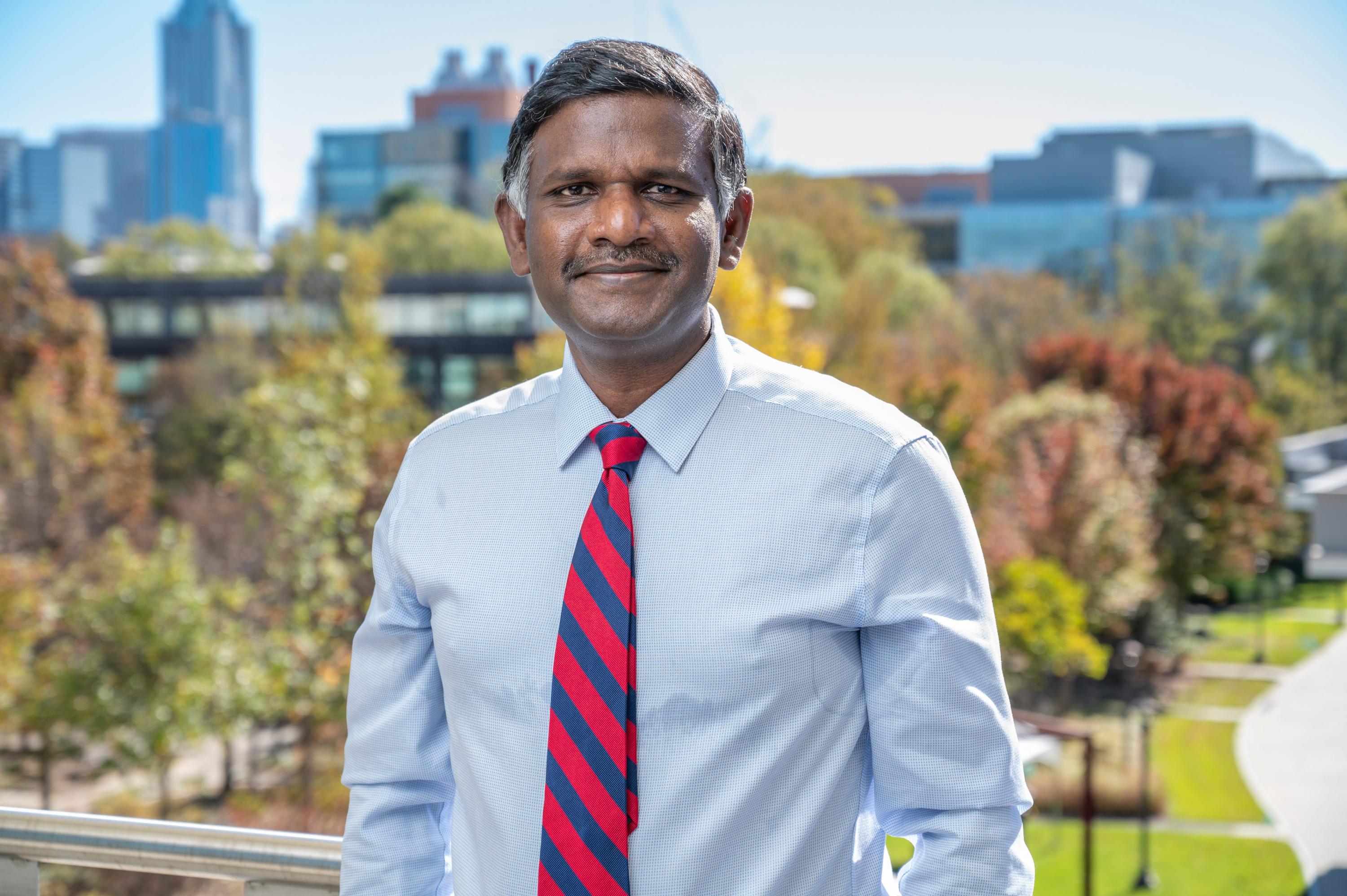 Photo of a man of south Asian descent with short dark hair and a smile. He is wearing a light blue button-down shirt and a red and royal blue striped tie. Behind him is the Atlanta skyline.