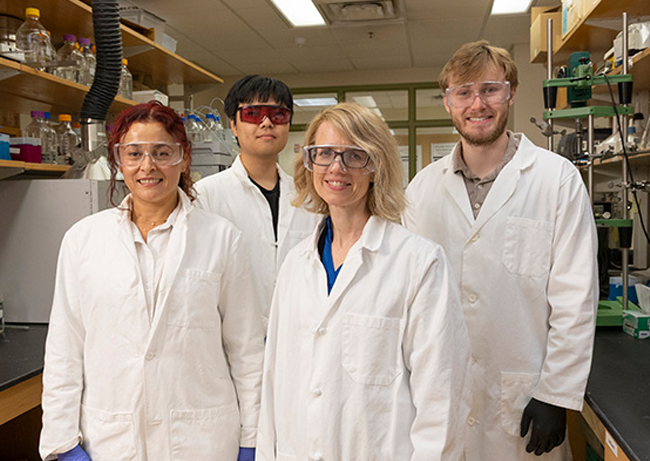 Four individuals wearing white lab coats stand together in a laboratory setting.