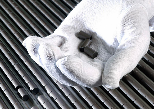 White protective glove holding several small cylindrical nuclear fuel pellets above rows of metal fuel rods.