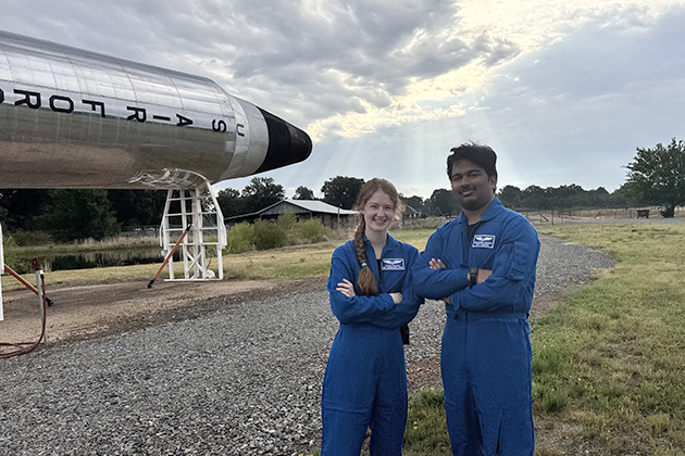 Two people wearing blue flight suits stand with arms crossed beside a large rocket display outdoors. Sunlight streams through clouds in the background above the grassy field and gravel path.