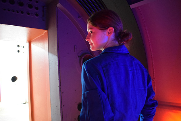 A person in a blue flight suit stands inside a curved, spacecraft-like corridor illuminated by blue and red lighting. The person is turned toward a brightly lit doorway at the end of the passage.
