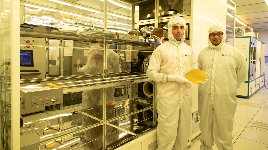 Two individuals wearing cleanroom suits stand inside a semiconductor manufacturing facility. One person holds a large silicon wafer while automated production equipment surrounds them.