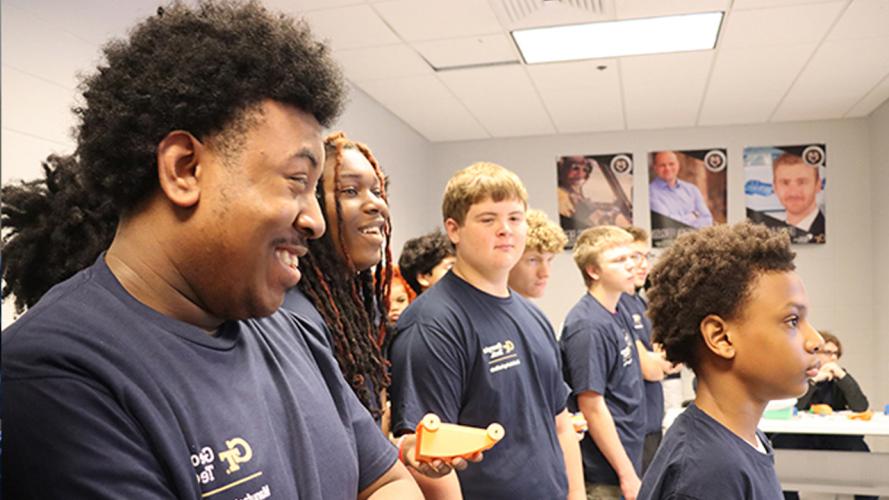 Group of students in navy shirts standing in a classroom, each holding small orange and blue 3D-printed objects.