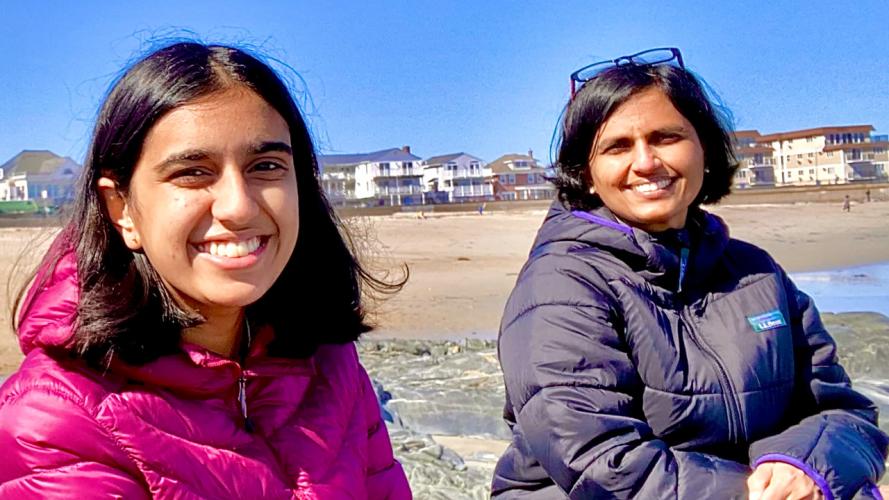 Two smiling researchers wearing jackets sit on rocks at a sunny beach with shoreline homes and the ocean in the background.