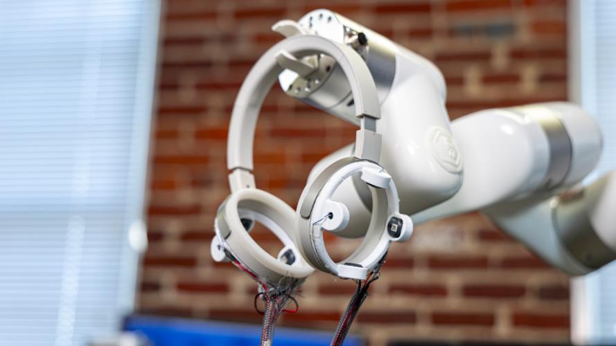 Robotic arm holding circular sensor devices with exposed wiring in a lab setting with a blurred brick wall background.