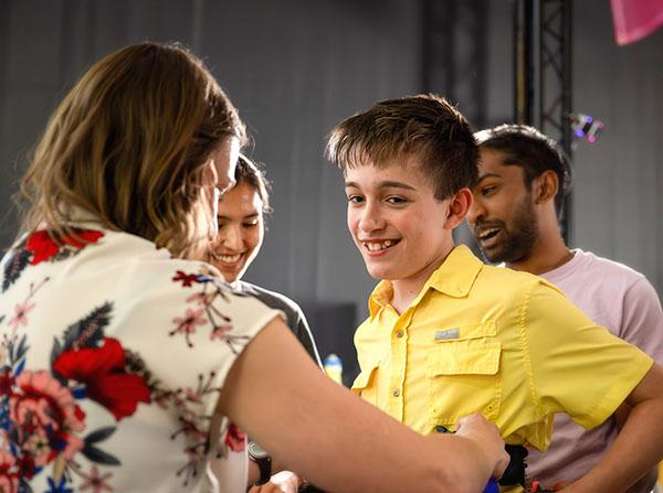Person wearing a floral-patterned shirt interacting with a group of people indoors; one individual is dressed in a bright yellow button-up shirt.