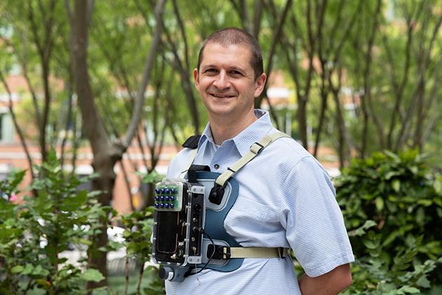 Man wearing a piece of equipment around his neck while standing in front of green trees and foilage